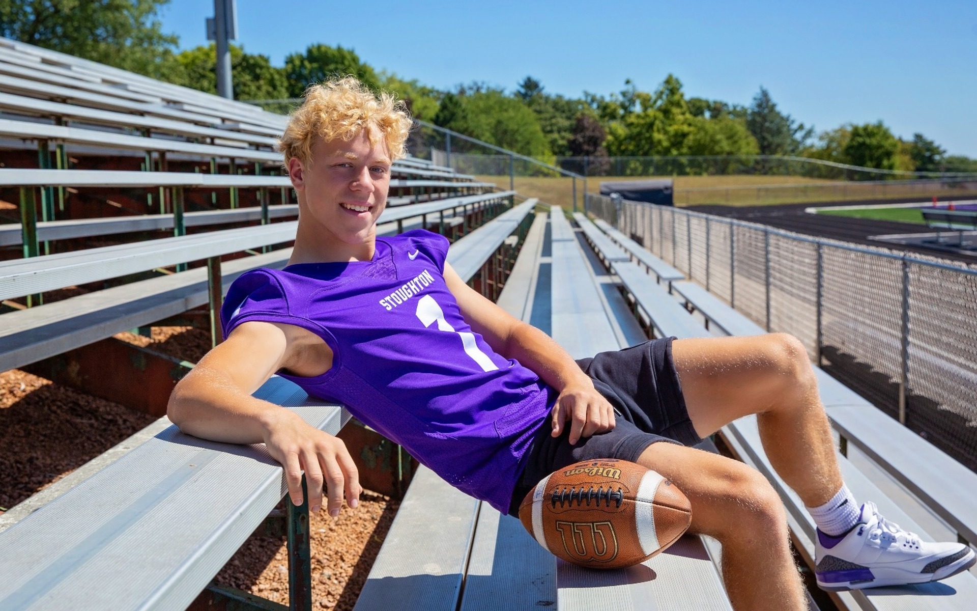 hs senior boy in a purple football jersey leans back on this bleachers field side