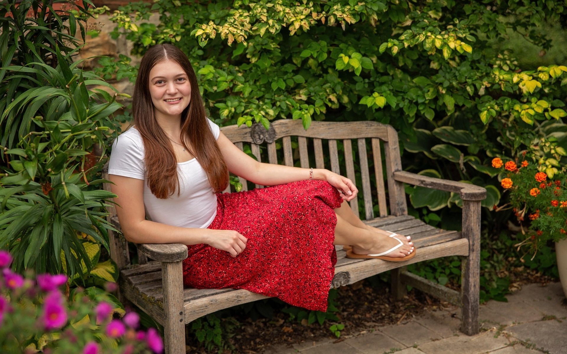 hs senior girl in red skirt sits on a park bench in a flower garden