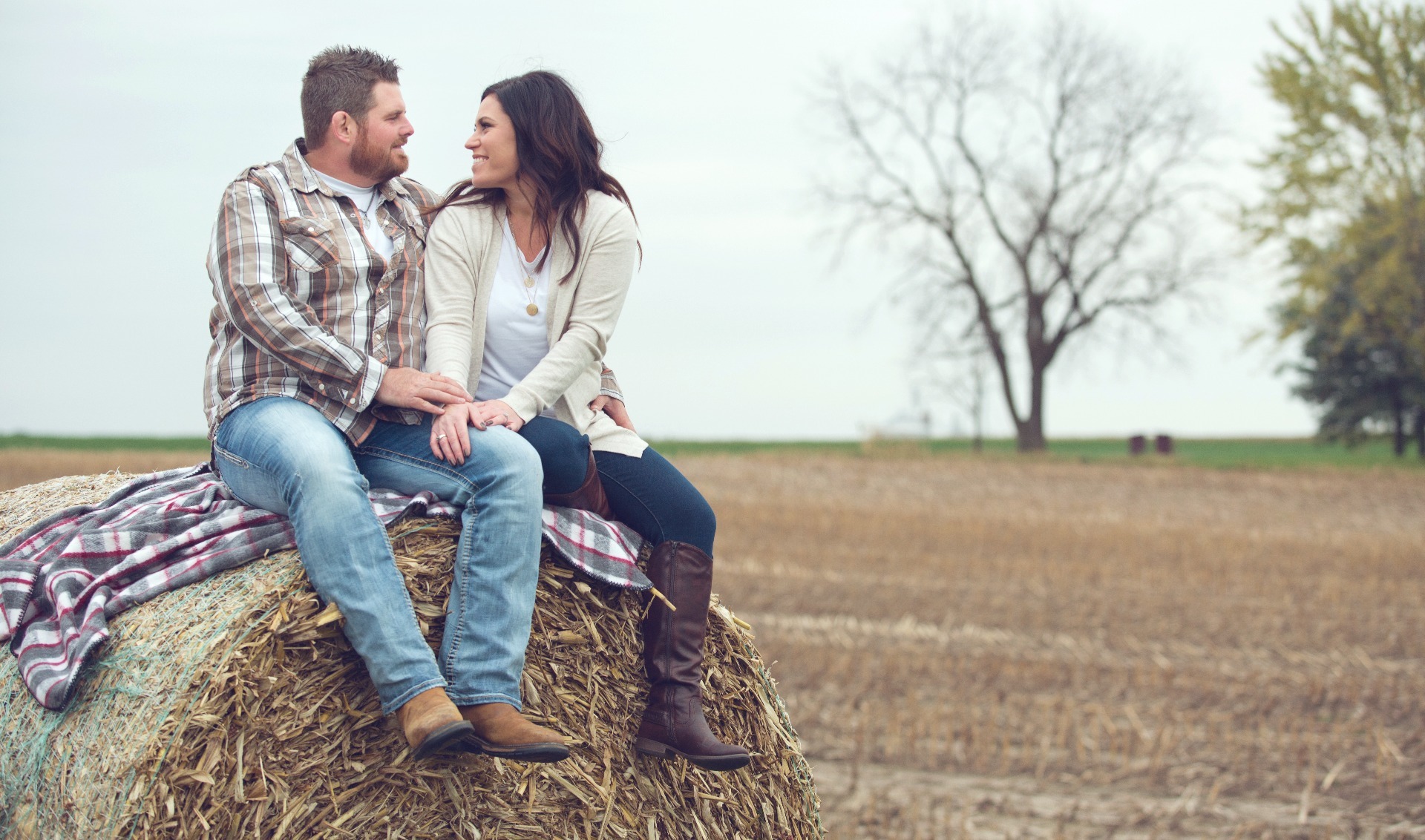 engaged couple sit atop a hale bale in a field