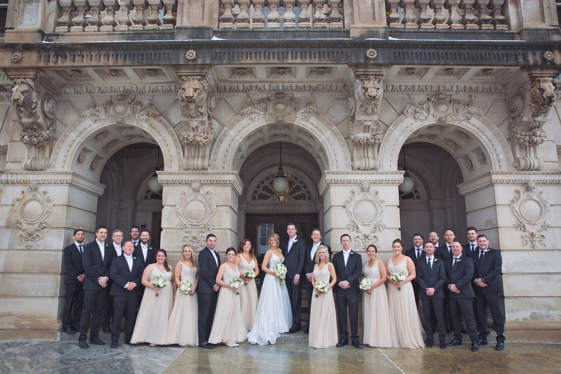 bride & groom pose with their large wedding party in front of a stone arched building