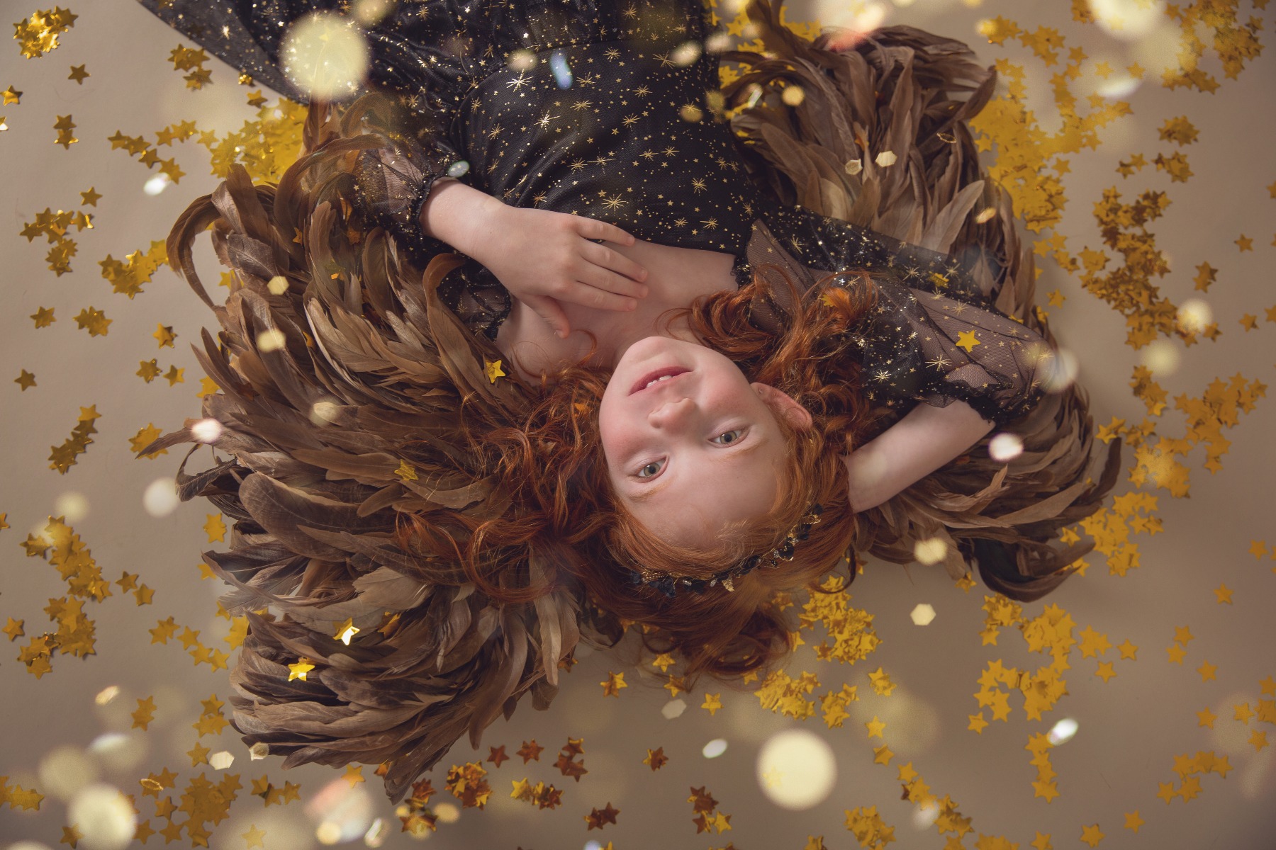 red headed girl with brown angel wings lays amongst a shower of gold confetti