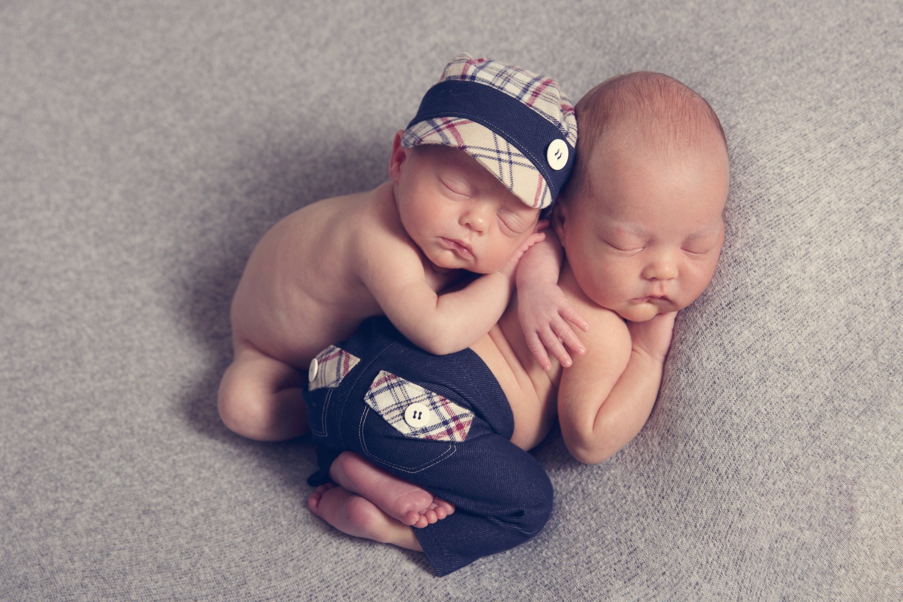 newborn twins sleeping on one another dressed in plaid hat & pants