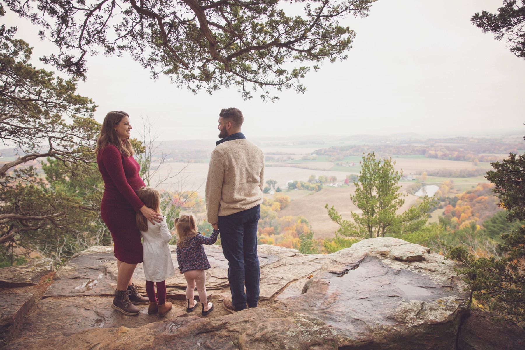 expectant mom, dad and two girls pose silhouette on a cliffside