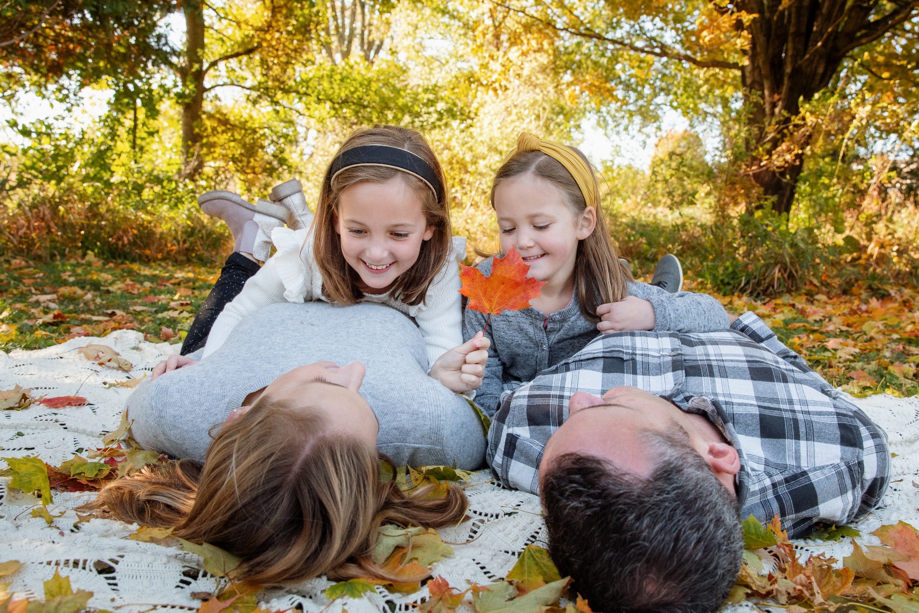 two young girls lay on their parents chest playing in the fall leaves