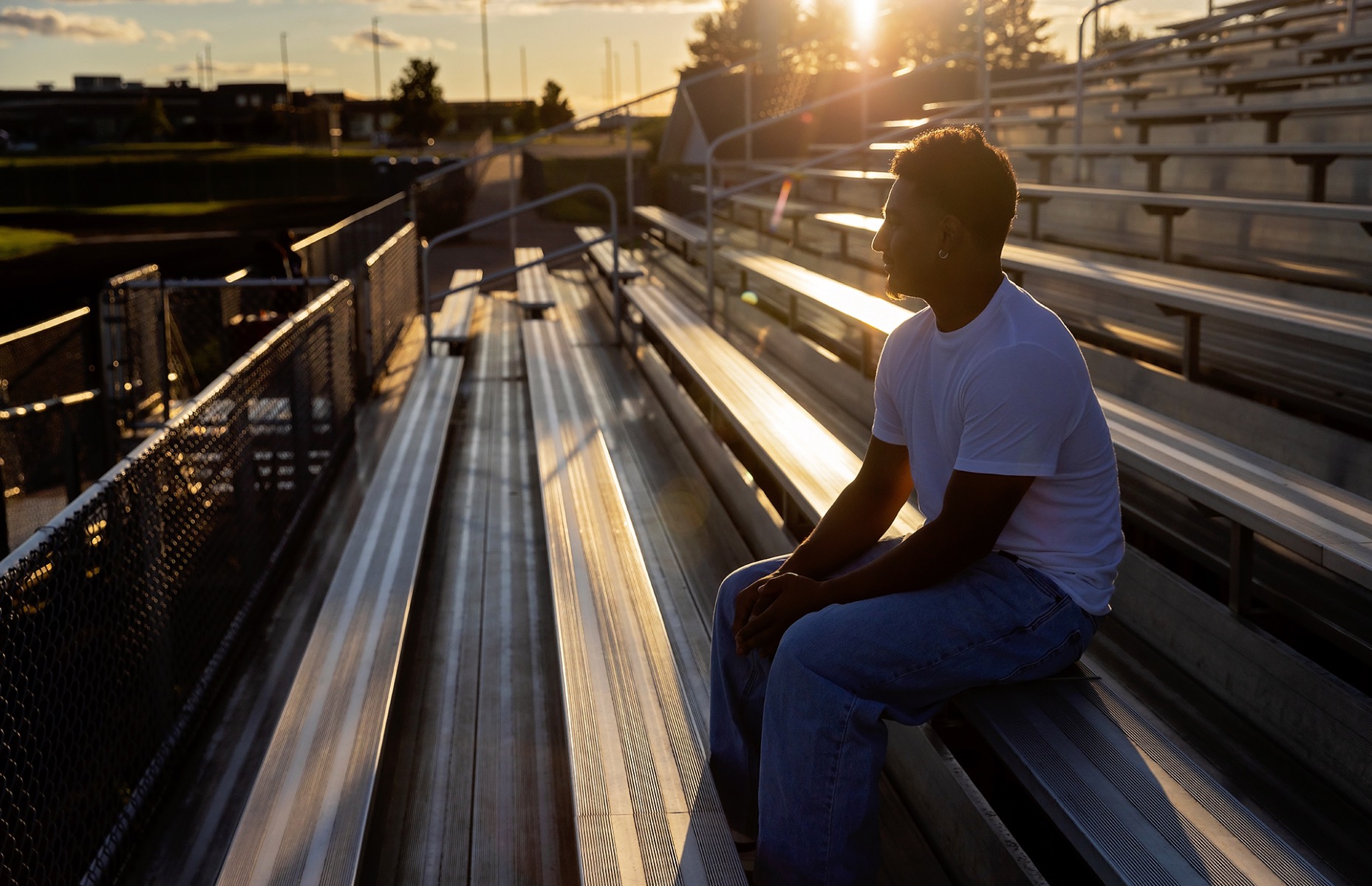 high school senior boy sits in silhouette on the bleachers of his schools football field